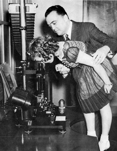 J. Edgar Hoover holds Shirley Temple as she peeks through a microscope at FBI headquarters.